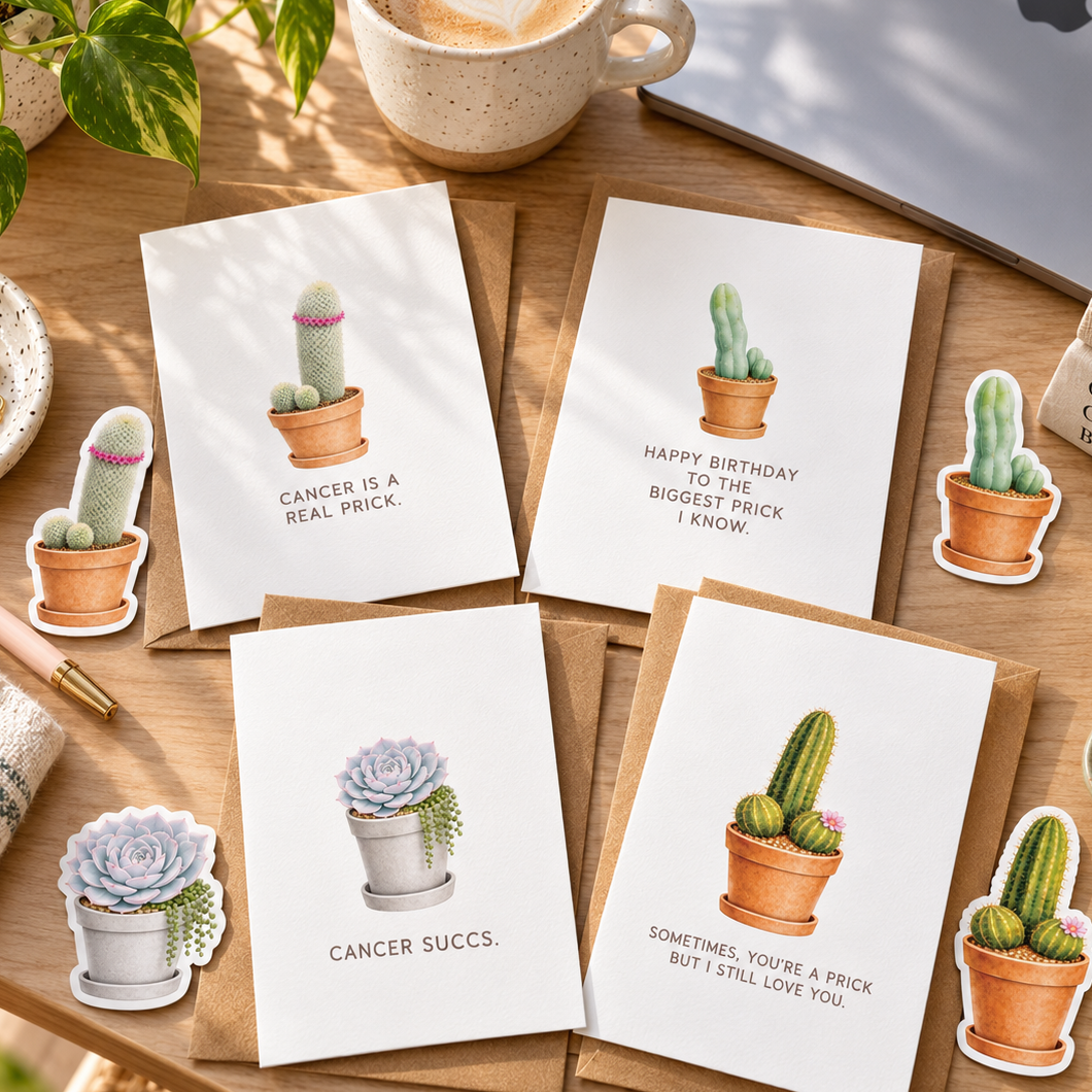 Four white greeting cards with cactus and succulent illustrations are arranged on a wooden desk with brown envelopes, a coffee cup, a laptop, a plant, and small decorative stickers. The cards have humorous cancer-themed quotes.