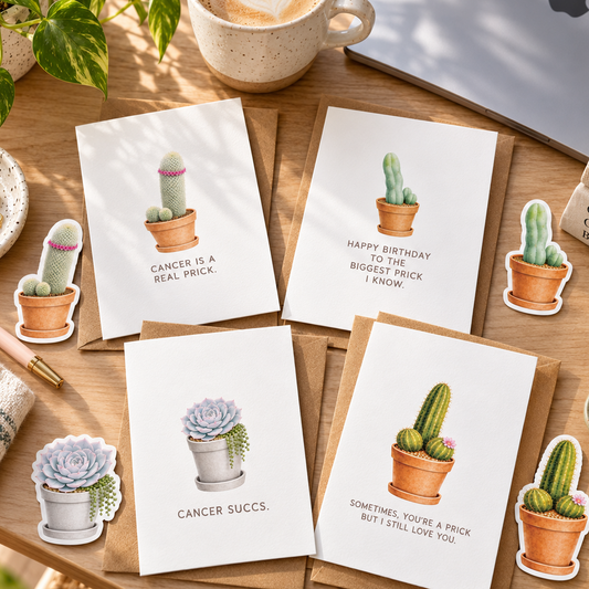 Four white greeting cards with cactus and succulent illustrations are arranged on a wooden desk with brown envelopes, a coffee cup, a laptop, a plant, and small decorative stickers. The cards have humorous cancer-themed quotes.